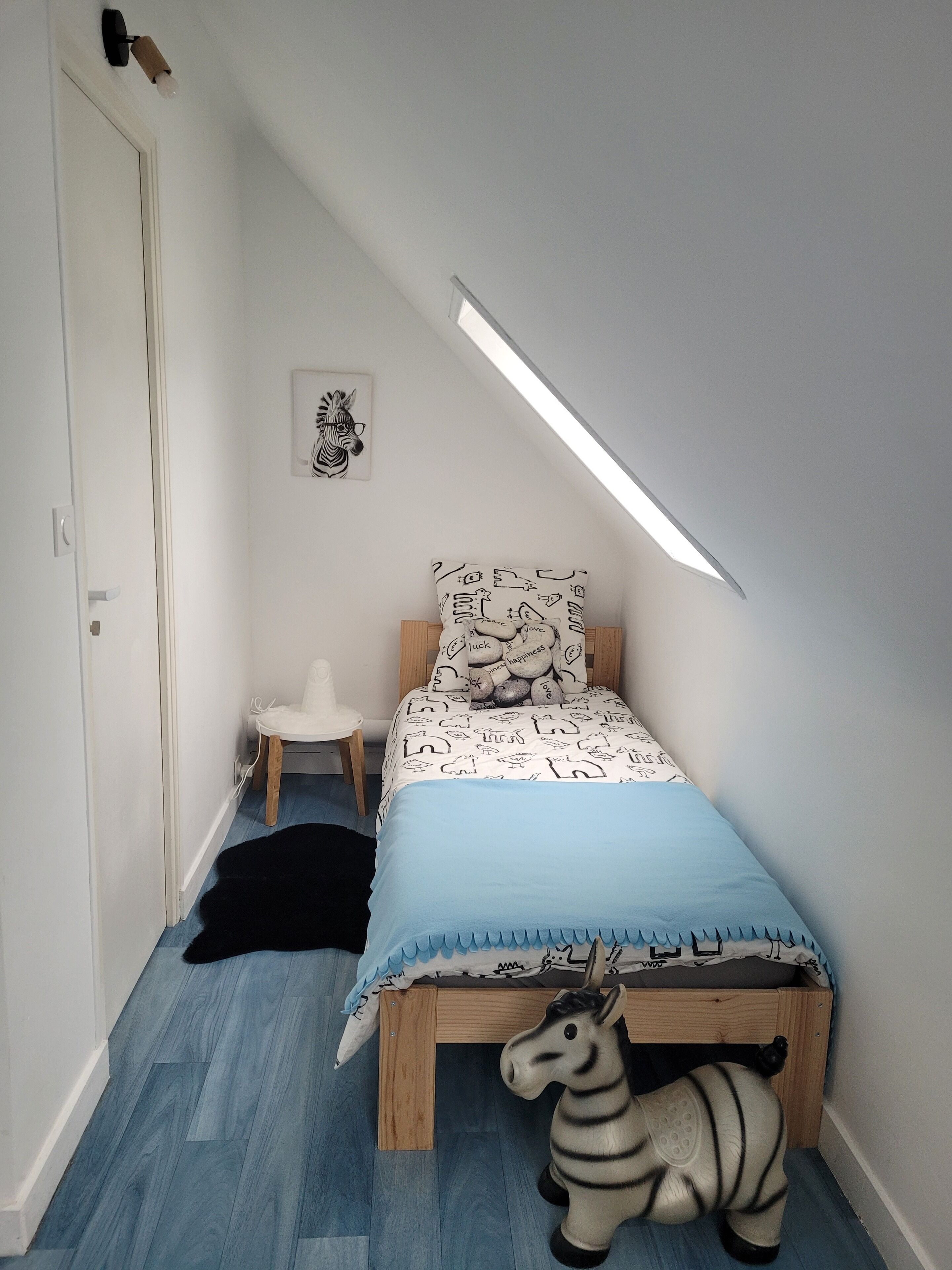 Ein Schlafzimmer mit schrägen Wänden, einem Bett mit gemusterten Betttextilien, einem Zebra-Spielzeug, einem kleinen Stuhl und einem an der Wand befestigten Zebra-Kopf als Kunstwerk.