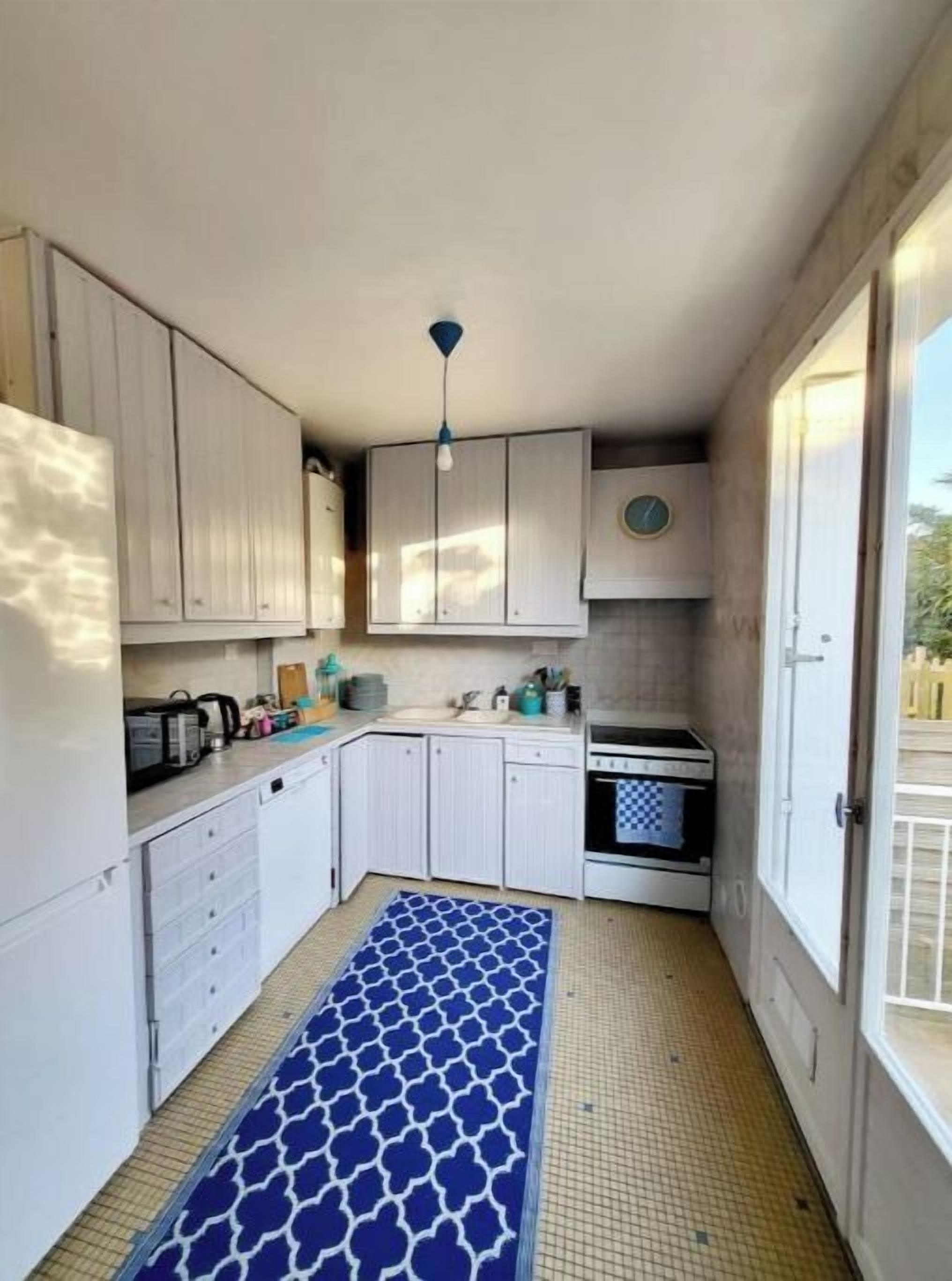 A compact kitchen with white cabinets, a blue patterned rug, and a view of the outside through a door.