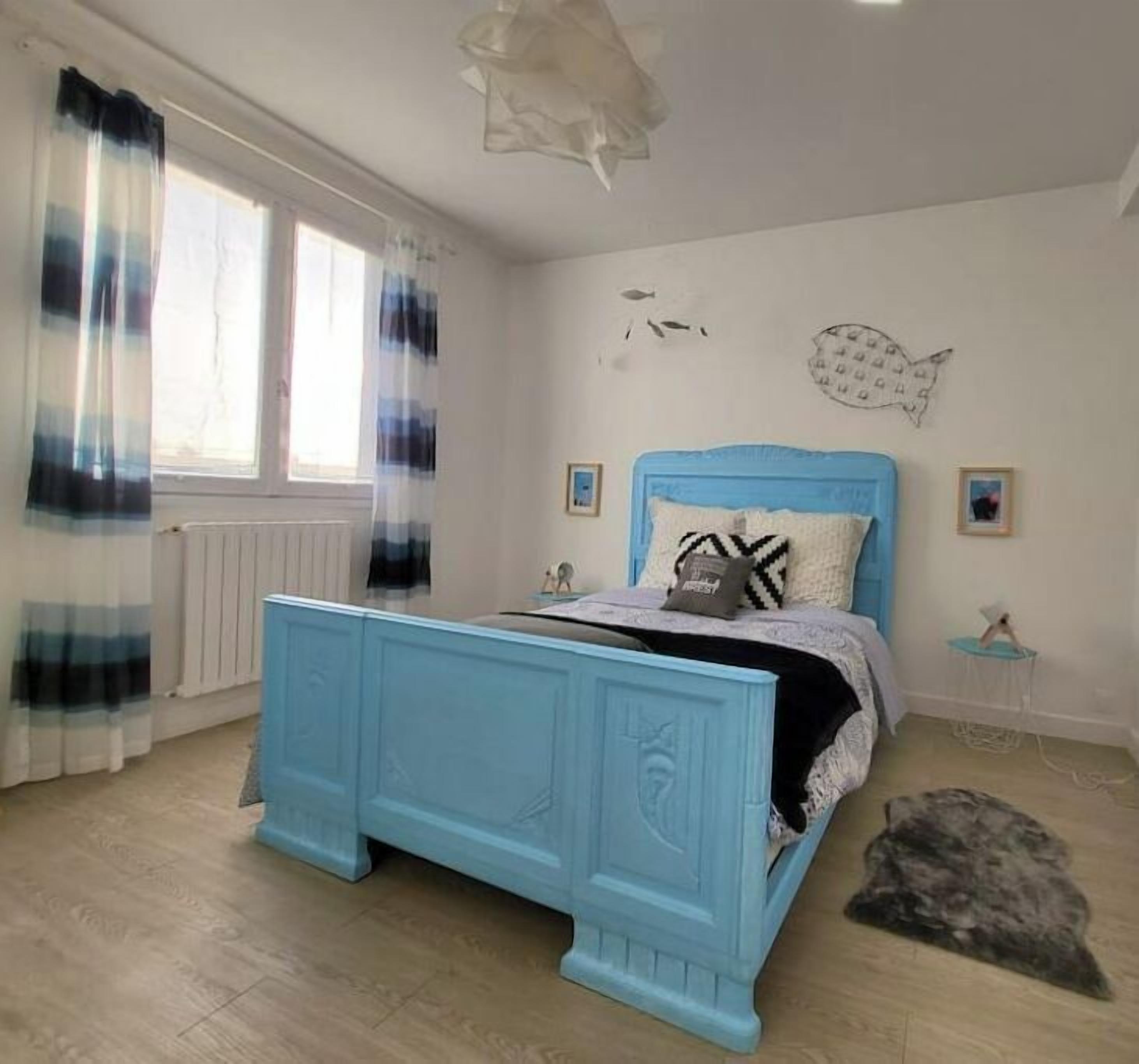 Een slaapkamer met een blauw bed, een raam met gordijnen, vis kunst aan de muur en een klein nachtkastje.