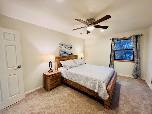 A bedroom with a wooden bed, two bedside tables with lamps, a ceiling fan, a window with curtains, and a door.
