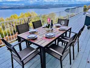 Eine Holzterrasse mit gedecktem Tisch für sechs Personen, mit Blick auf einen See und Berge.