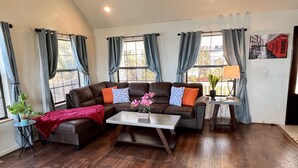 A living room with a brown leather sectional sofa, a coffee table, and a small side table with a lamp. There are two windows with curtains, a potted plant, and a wall-mounted picture. - Amazing Lake View Home w Pool table Ping-Pong BBQ Deck, Watch Sunset  (Kelseyville)