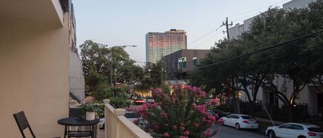 A balcony with a table and chairs, a potted plant, and a view of a street with cars and buildings.