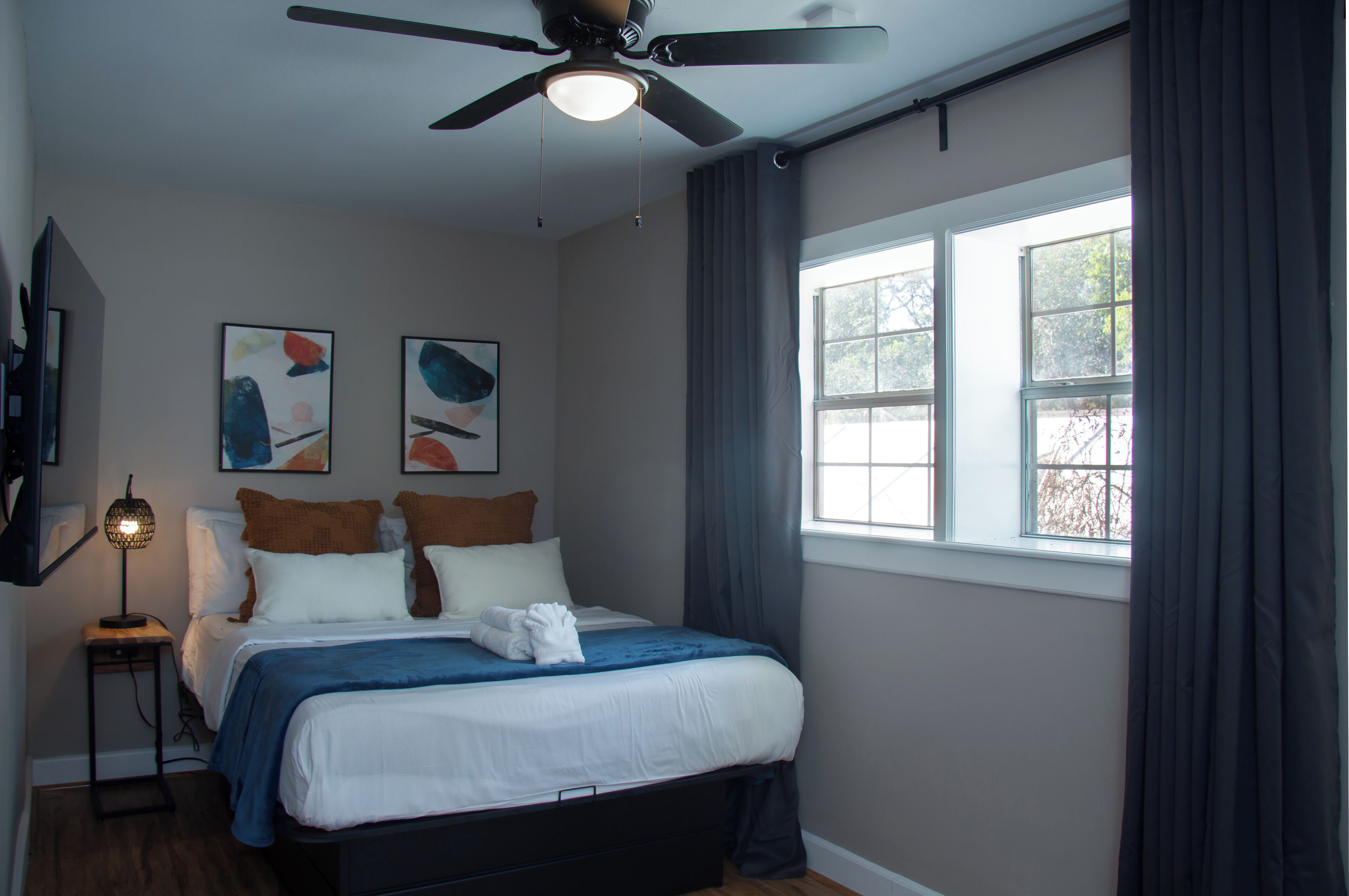 Un dormitorio con una cama, ventilador de techo, ventana con cortinas y obras de arte en la pared.