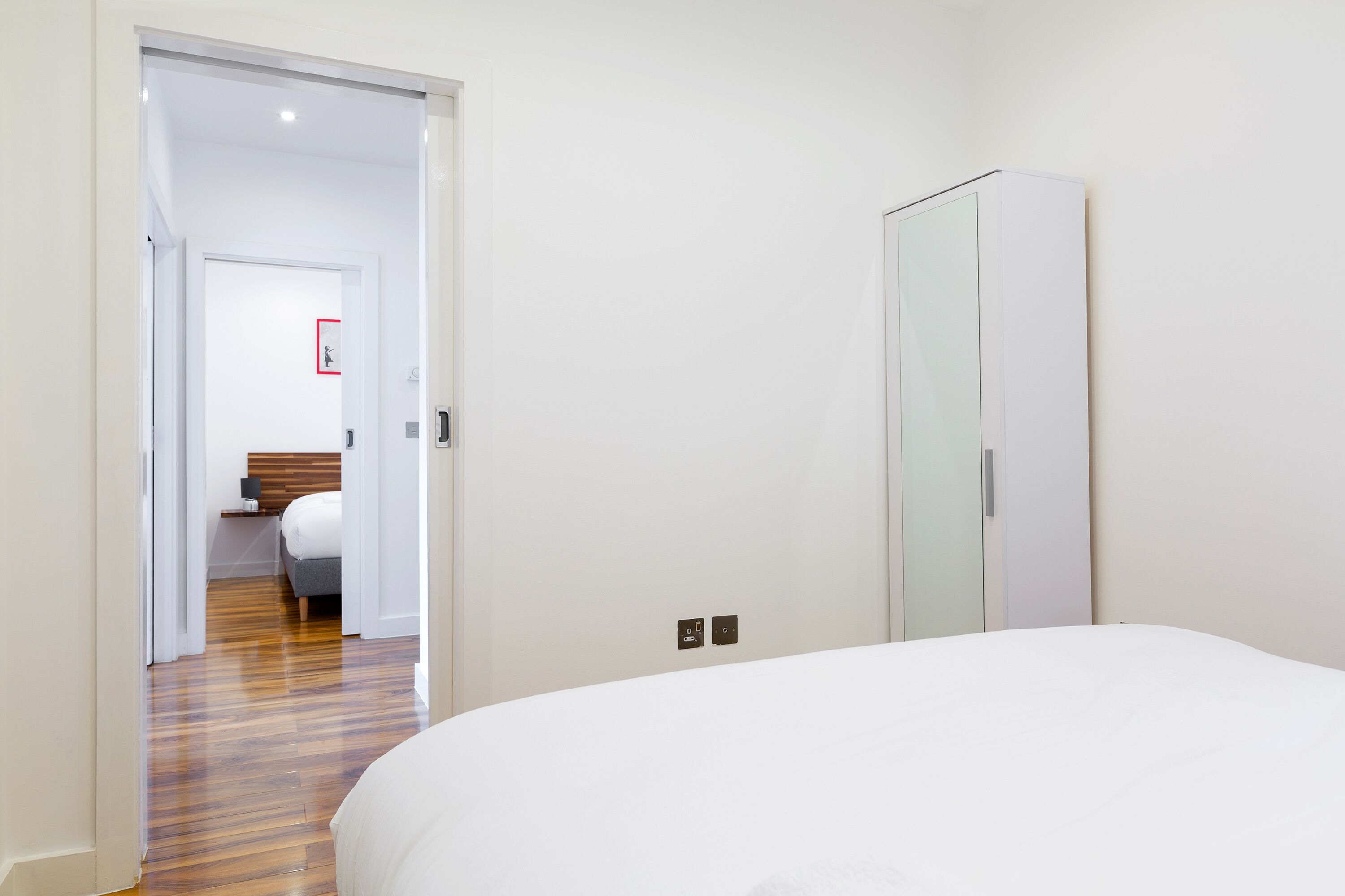A hotel room with a wooden floor, a white bed, a wardrobe, and a door leading to a bathroom with a mirror and a towel rack.