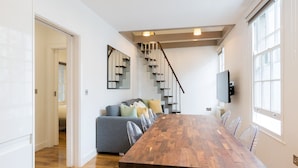 A modern interior with a wooden dining table, clear plastic chairs, a grey sofa with cushions, a staircase, and a TV mounted on the wall.