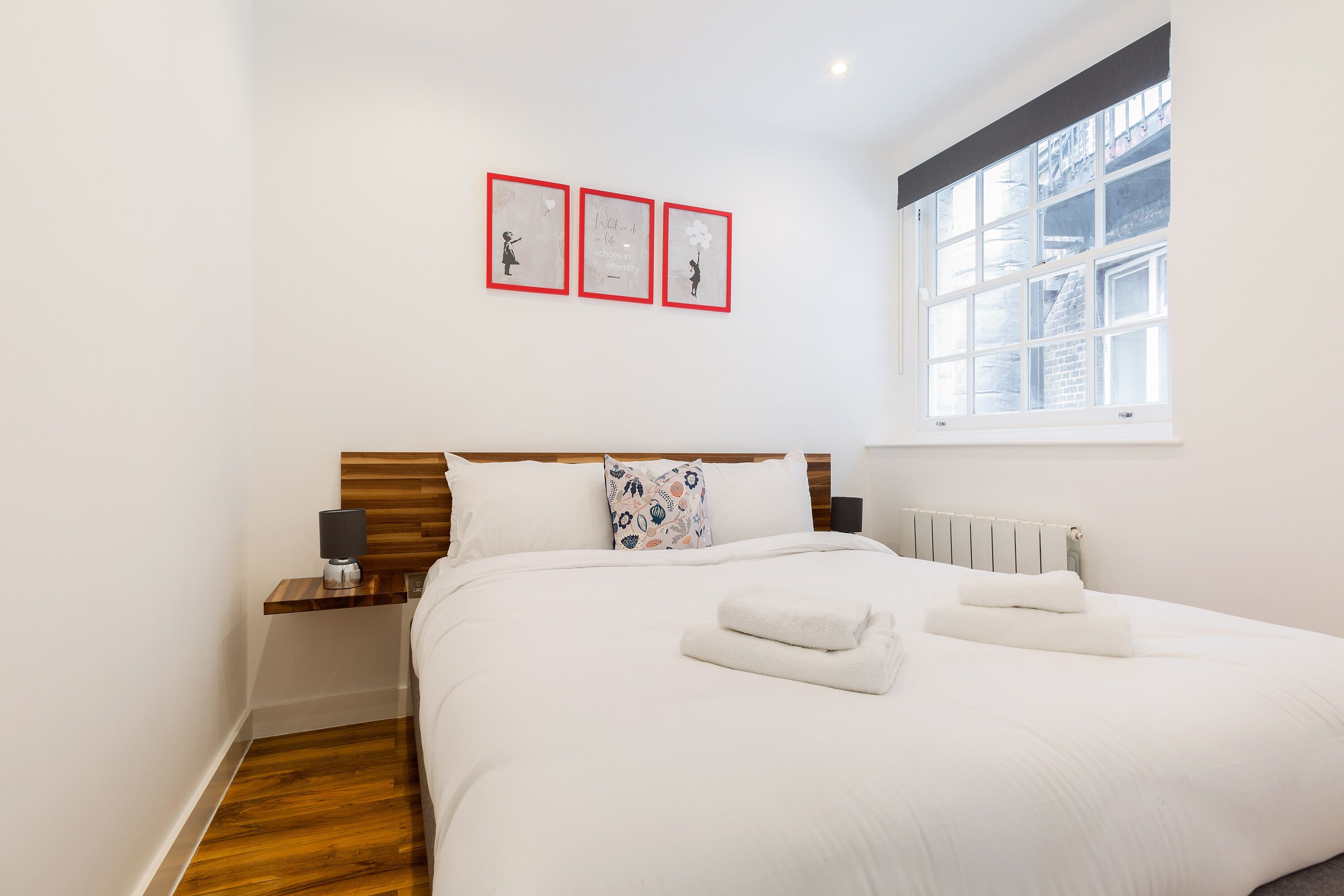 A neatly made bed with white linens and two folded towels, a wooden headboard, a bedside table with a lamp, and a window with a view of buildings.
