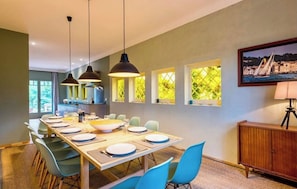 A dining area with a long table set for six, blue chairs, pendant lights, and a view of the sea through windows.