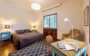 Un dormitorio con una cama, dos mesitas de noche con lámparas, una silla, una mesa con revistas, una ventana con persianas y una lámpara en el techo.