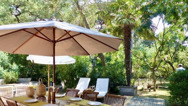 Ein Essbereich im Freien mit einem Holztisch und -stühlen, einem großen Sonnenschirm und Blick auf einen Garten mit Bäumen und einer Holzterrasse.