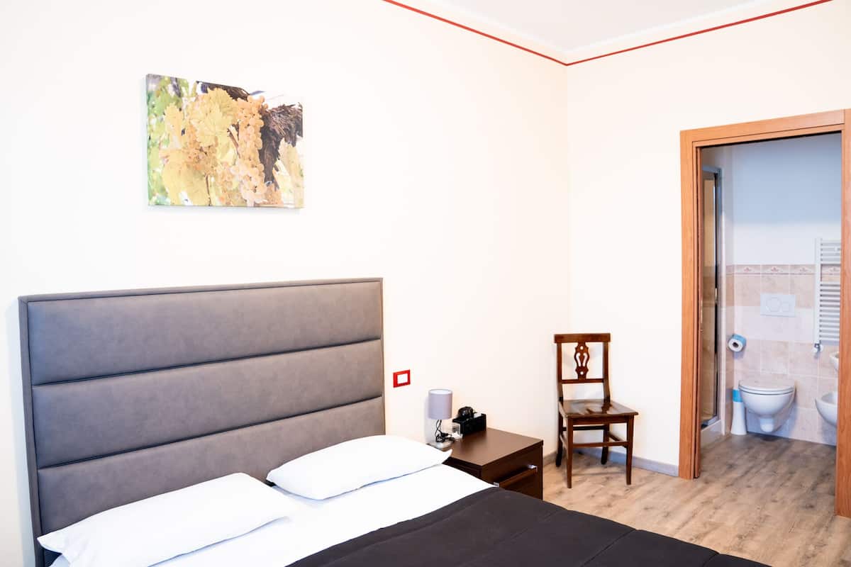 Un dormitorio con una cama grande, una silla, una mesita de noche y un baño visible a través de una puerta abierta.