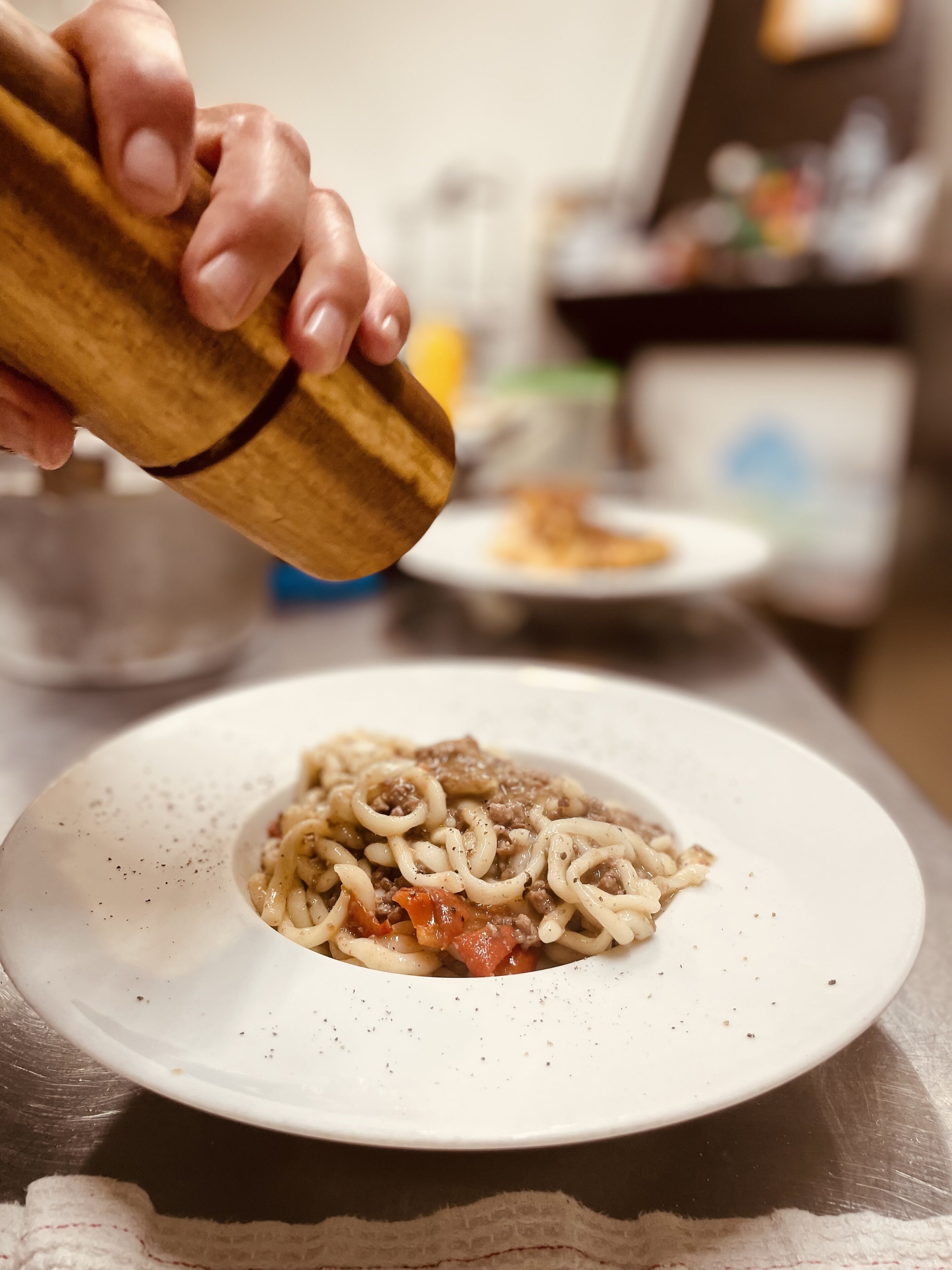 A hand holding a pepper grinder over a plate of pasta with meat and tomato sauce.