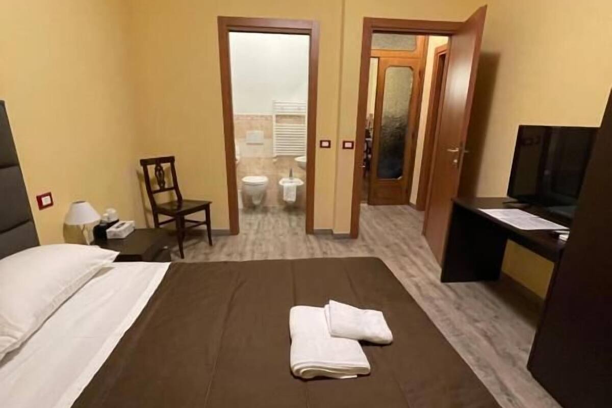 Habitación de hotel con cama, silla, escritorio, televisor y baño visible a través de una puerta abierta.