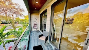 Een balkon met glazen hekwerk, buitenmeubilair en uitzicht op een tuin en bomen.