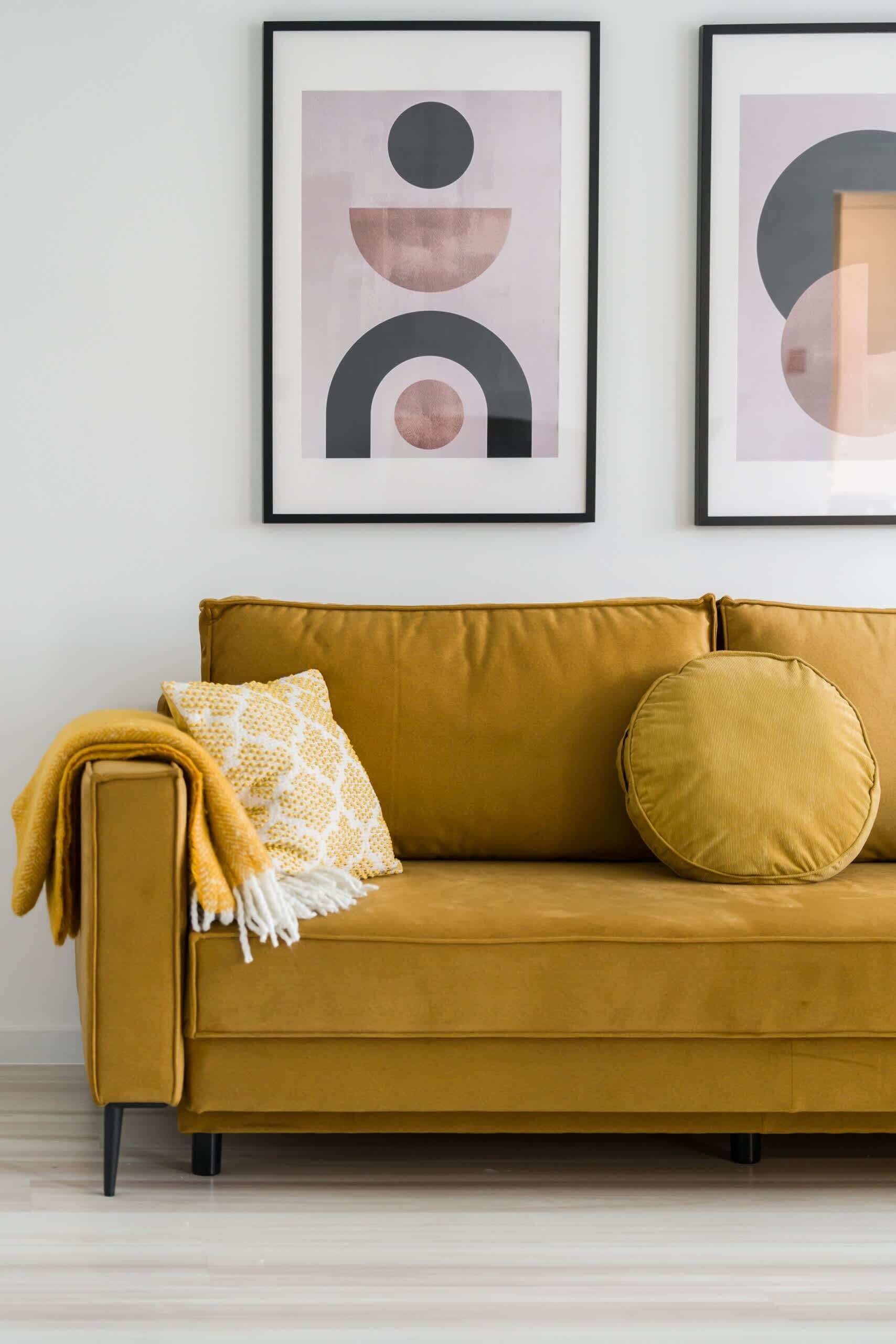 Un sofá color mostaza con una manta estampada y dos cojines, frente a una pared con dos obras de arte abstracto.