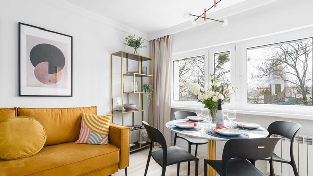 Un salon moderne avec un canapé de couleur moutarde, une bibliothèque avec un cadre doré et un coin repas comprenant une table ronde et des chaises noires.