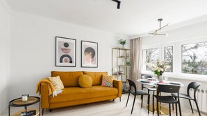 Un salon moderne avec un canapé de couleur moutarde, une table de salle à manger ronde entourée de chaises noires et une bibliothèque avec un cadre doré.