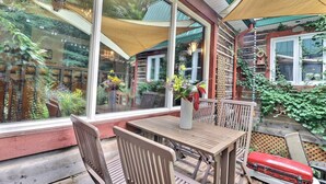 A wooden deck with a table and chairs, a red suitcase, and a large window overlooking a garden. - MA GALÈRE, Eco-responsible house by the river. (Saint-Michel-de-Bellechasse)
