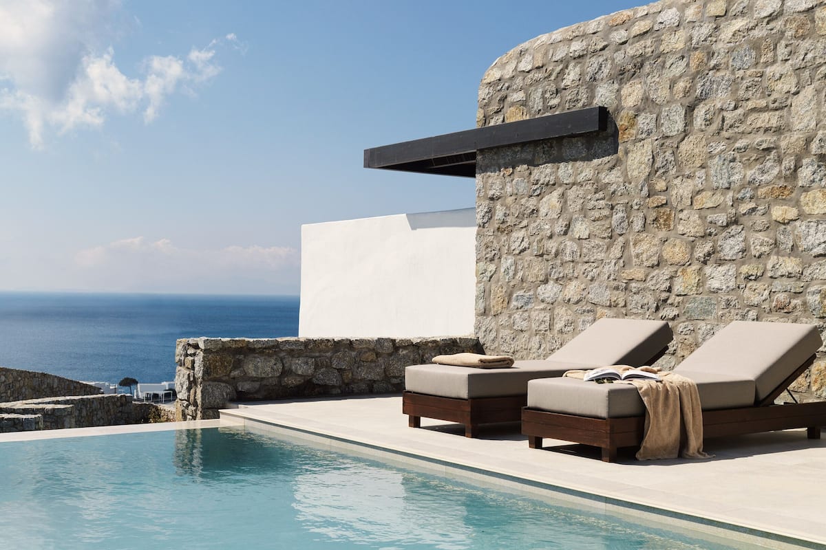 Un espace aménagé au bord de la piscine, avec des chaises longues et un mur en pierre, donnant sur une étendue d’eau.