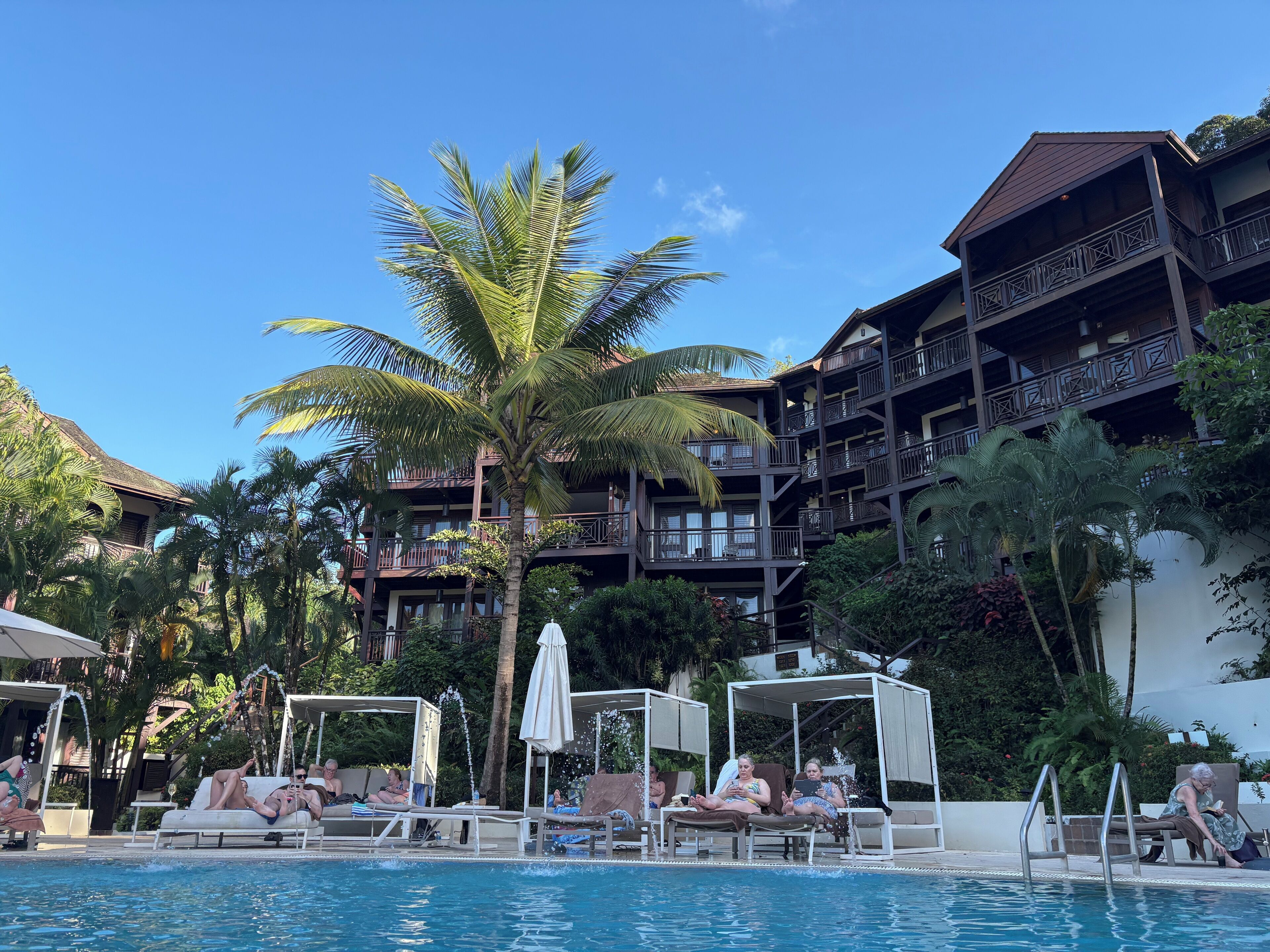 A pool area with lounge chairs and umbrellas, surrounded by palm trees and a multi-story building with balconies.
