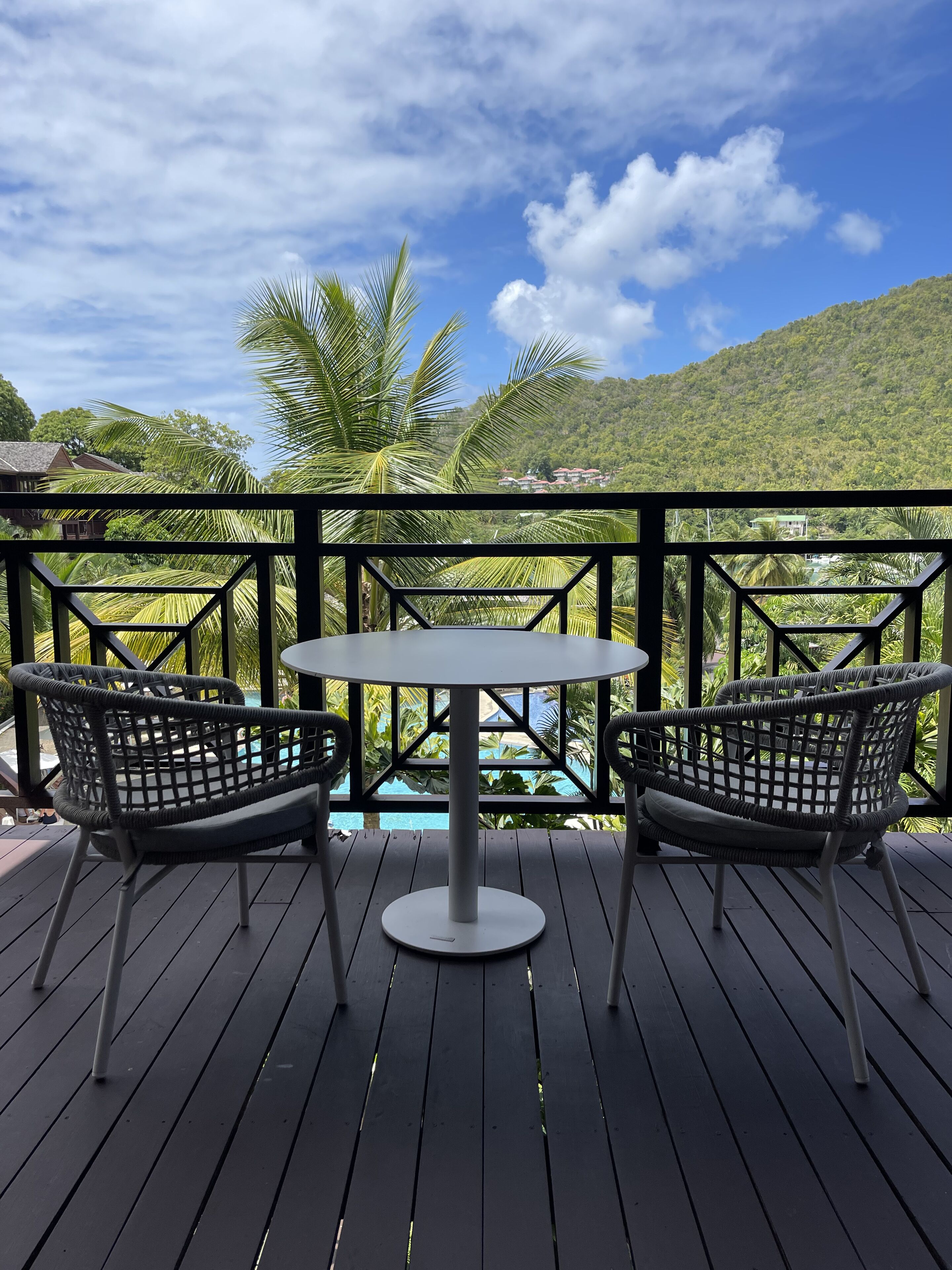 Eine Holzterrasse mit zwei Korbstühlen und einem runden Tisch, mit Blick auf einen Pool und üppiges Grün.