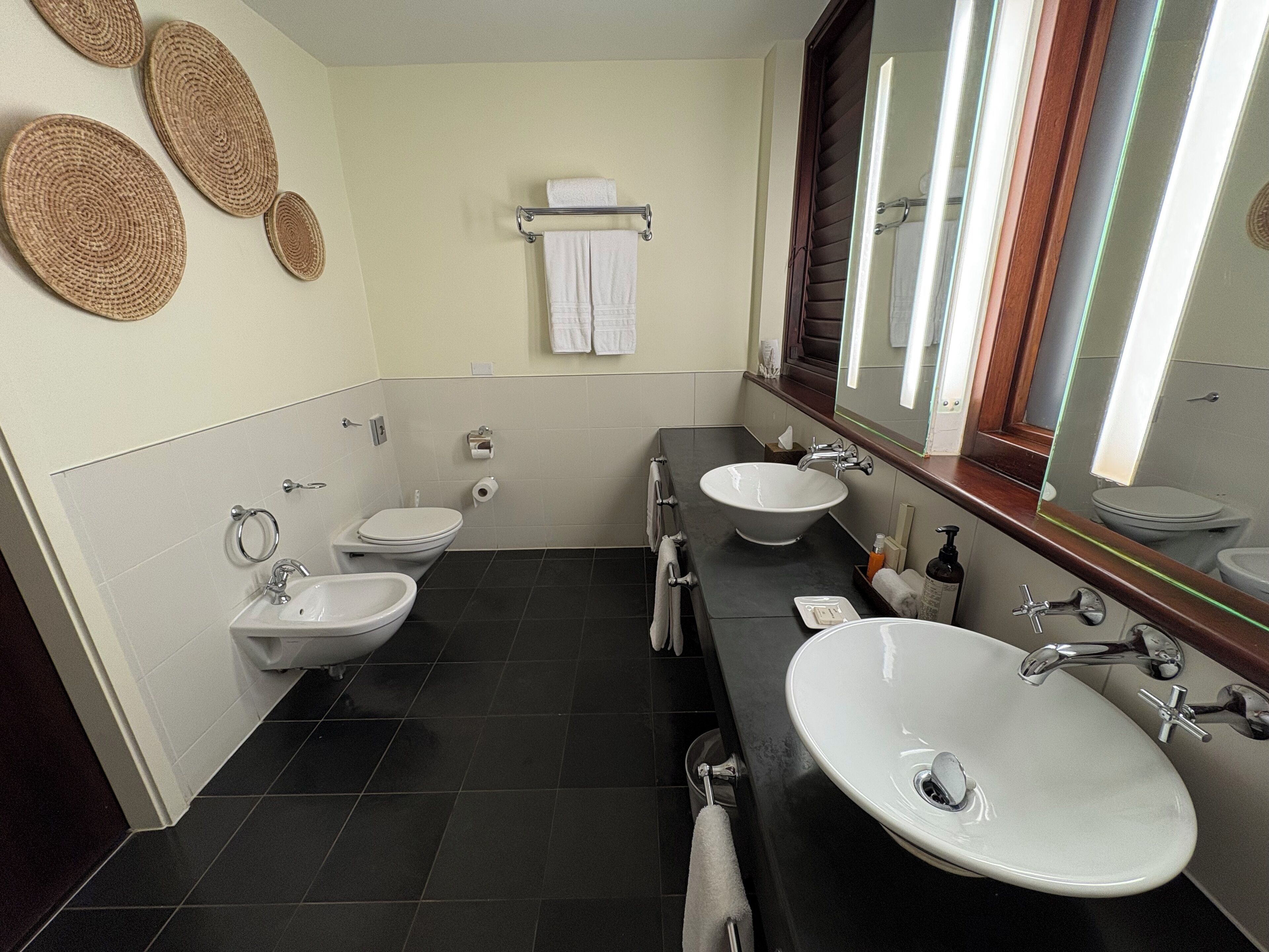 Ein Badezimmer mit zwei Waschbecken, einer Toilette und einem Spiegel. Handtücher hängen an der Wand und ein Ablagebereich enthält Toilettenartikel.