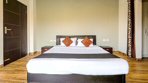 A large bed with white bedding and two decorative pillows, flanked by nightstands with lamps, in a room with wooden flooring and a dark door. - IVA 1 BHK - w/ Wi-Fi, Kitchen, Food, Balcony, Garden (Sukha)