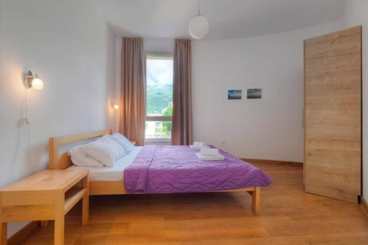 A room with a bed, a wooden nightstand, a window with curtains, and a wooden door.