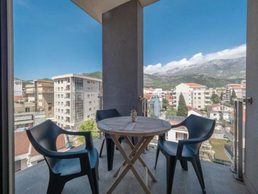 Un balcon avec une table ronde et deux chaises, donnant sur un paysage urbain avec des immeubles et des montagnes.