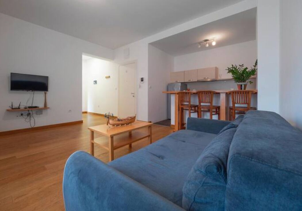 A modern living room with a blue sofa, wooden dining table, and a kitchen area with cabinets and a TV.