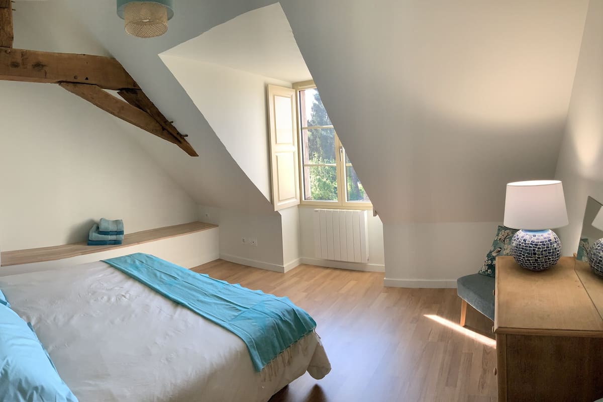 Une chambre avec un plafond en pente, un lit avec une literie bleue, une table de chevet avec une lampe et une fenêtre donnant sur un paysage verdoyant.