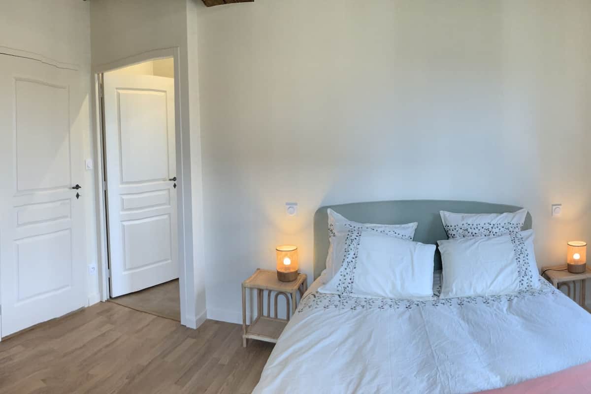 Une chambre à coucher avec un lit, deux tables de chevet avec des lampes, un fauteuil en osier et une fenêtre avec des rideaux.