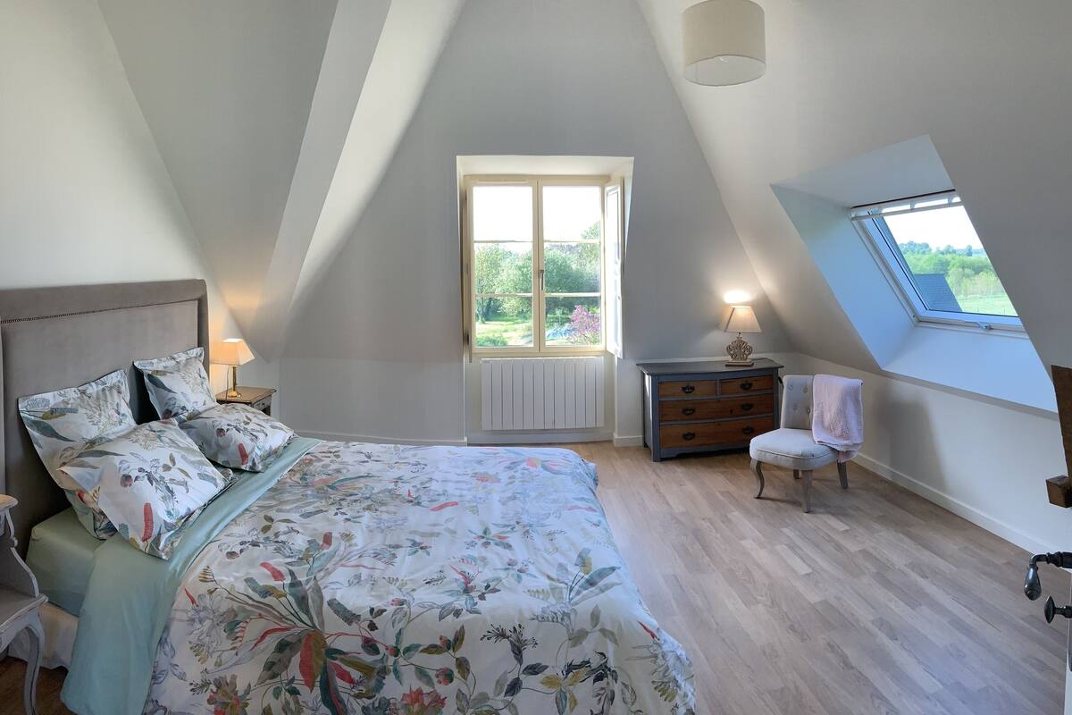 Une chambre avec un plafond en pente, un grand lit avec une literie à motifs floraux, une commode, une chaise et une fenêtre donnant sur un paysage verdoyant.