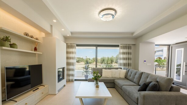 A modern living room with a flat-screen TV, a sofa, a coffee table, and a large window offering a view of greenery. - Violet Maison (Kos)