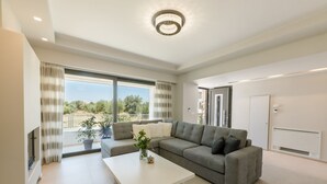 A modern living room with a grey sectional sofa, a white coffee table, and a large window offering a view of greenery and a building. - Violet Maison (Kos)