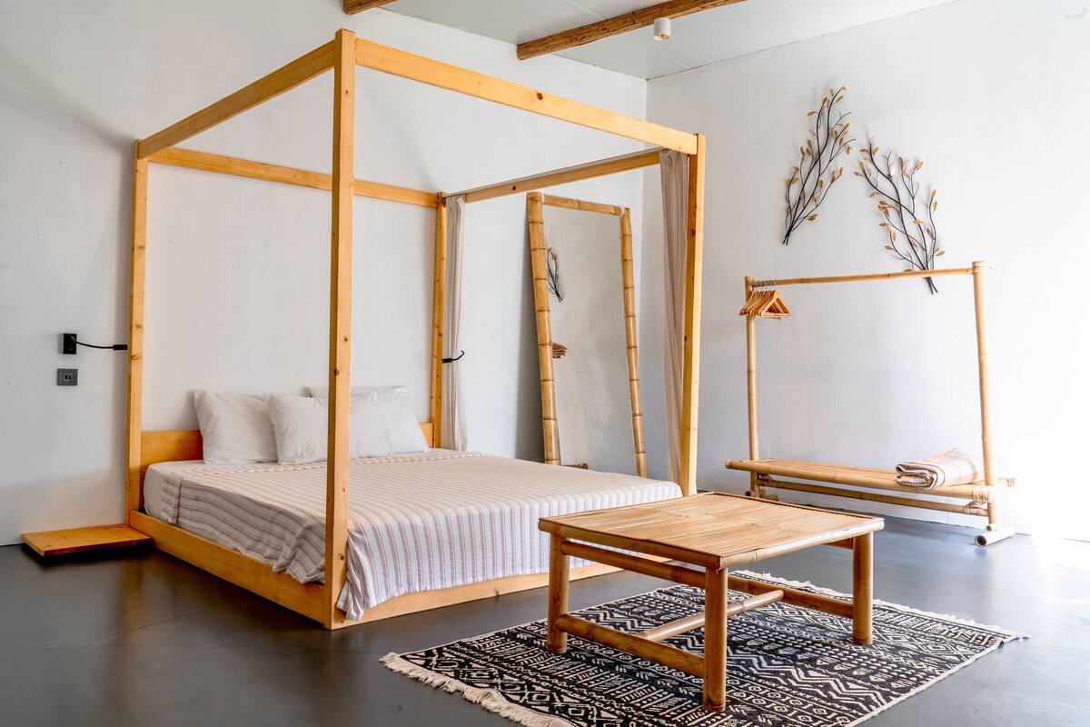 Una cama con dosel de madera, una mesita pequeña y un tapete estampado en una habitación minimalista con suelo de concreto y paredes blancas.