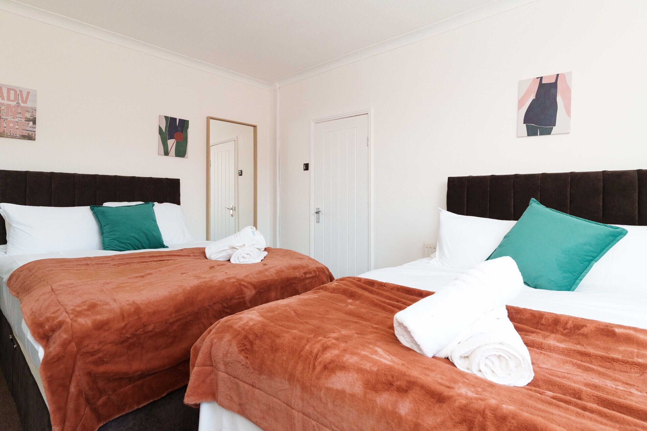 A hotel room with two beds, each with a brown headboard, white bedding, and teal pillows. There is a white door and a small artwork on the wall.