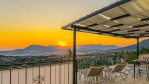 Un balcon offrant une vue sur le coucher de soleil derrière les montagnes, aménagé avec deux chaises et une table.