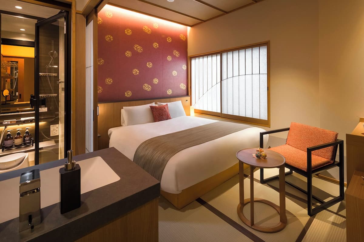 A hotel room with a bed, a chair, a small table, a window with a view, and a kitchenette area.