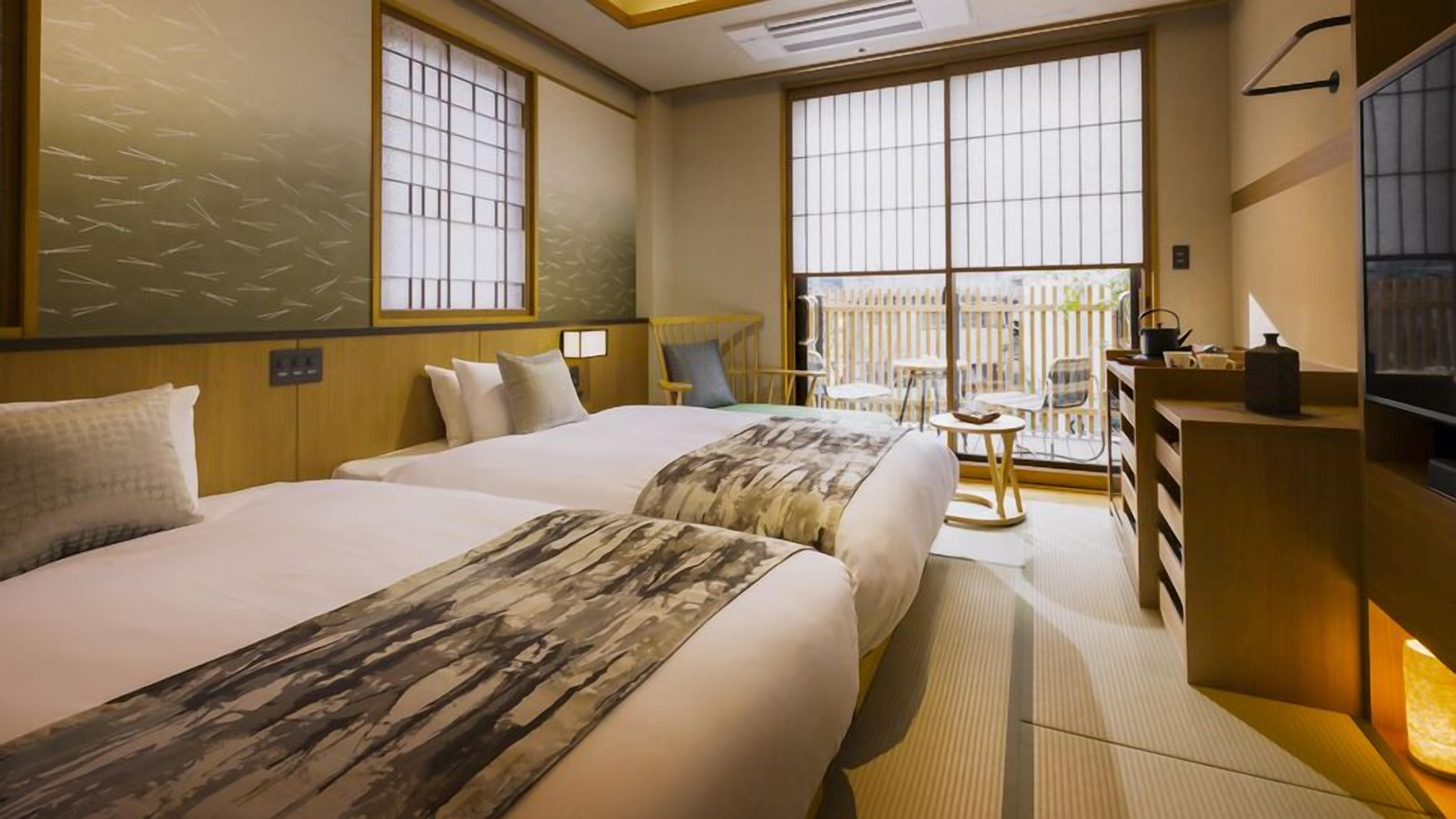 A hotel room with two beds, a desk, a chair, and large windows.