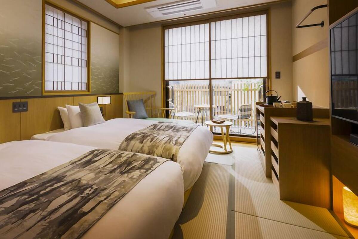 A hotel room with two beds, a desk, a chair, and large windows.