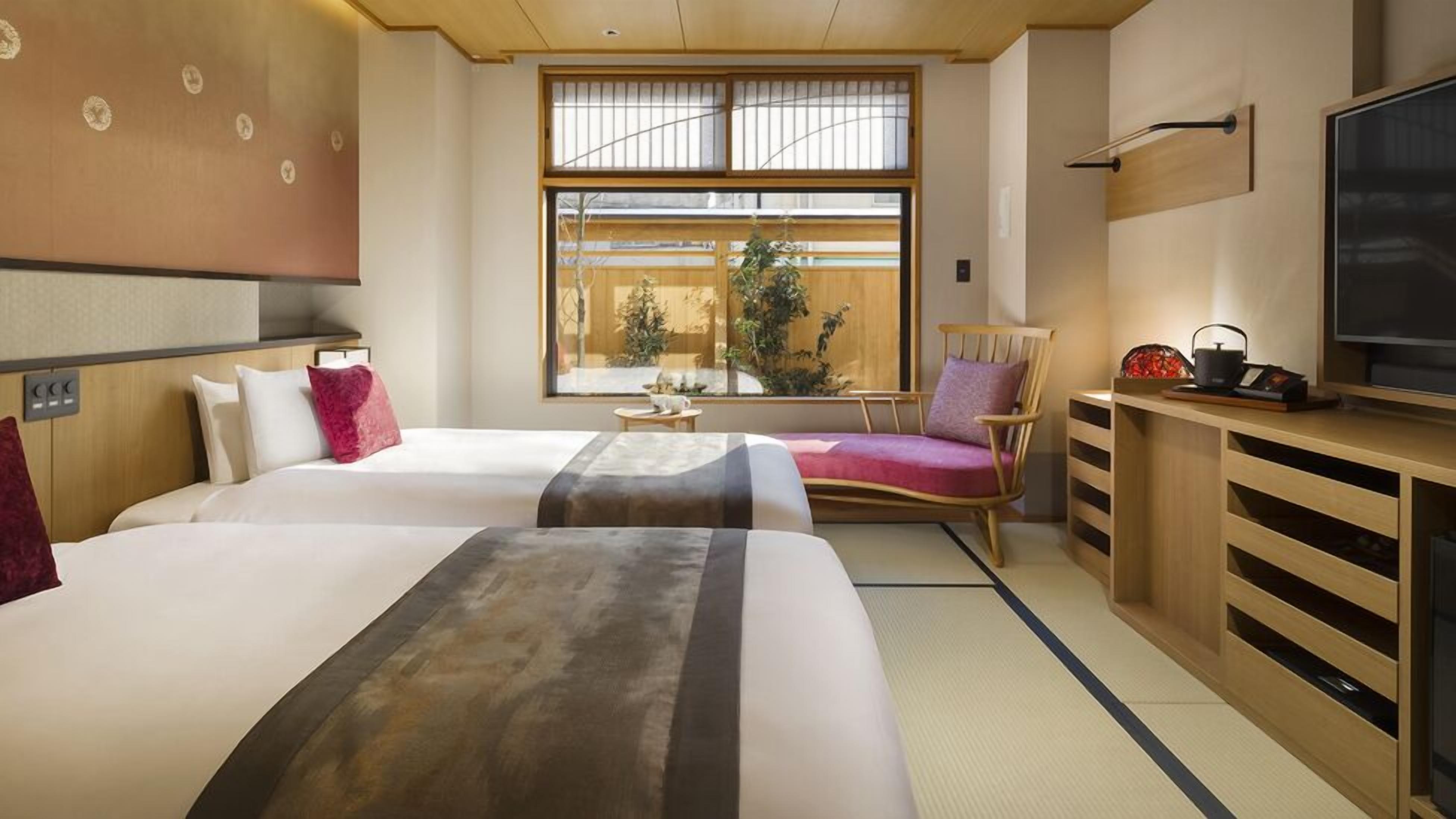A hotel room with two beds, a TV, a wooden desk, a chair, and a large window with a view of greenery.