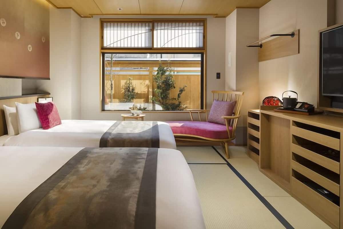 A hotel room with two beds, a TV, a wooden desk, a chair, and a large window with a view of greenery.