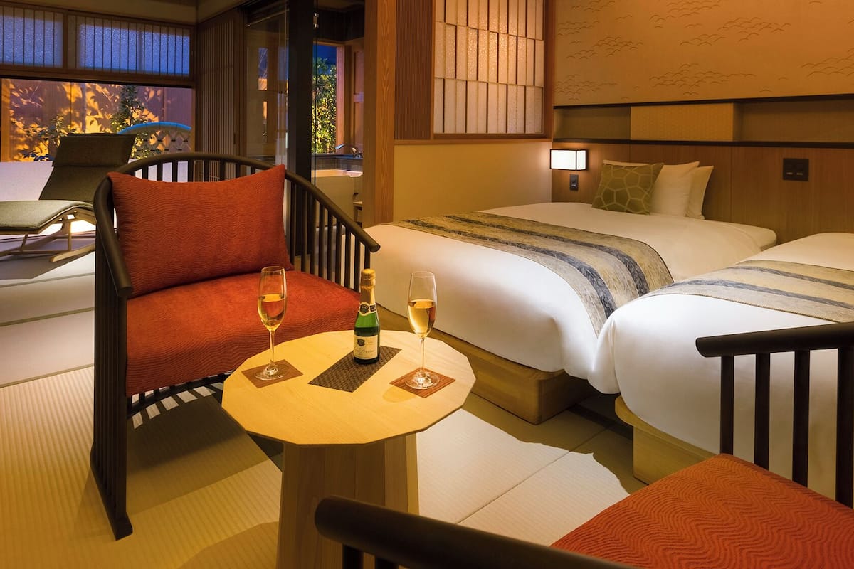 A hotel room with a sofa, two beds, a table with glasses and a bottle, and a view of the outside.