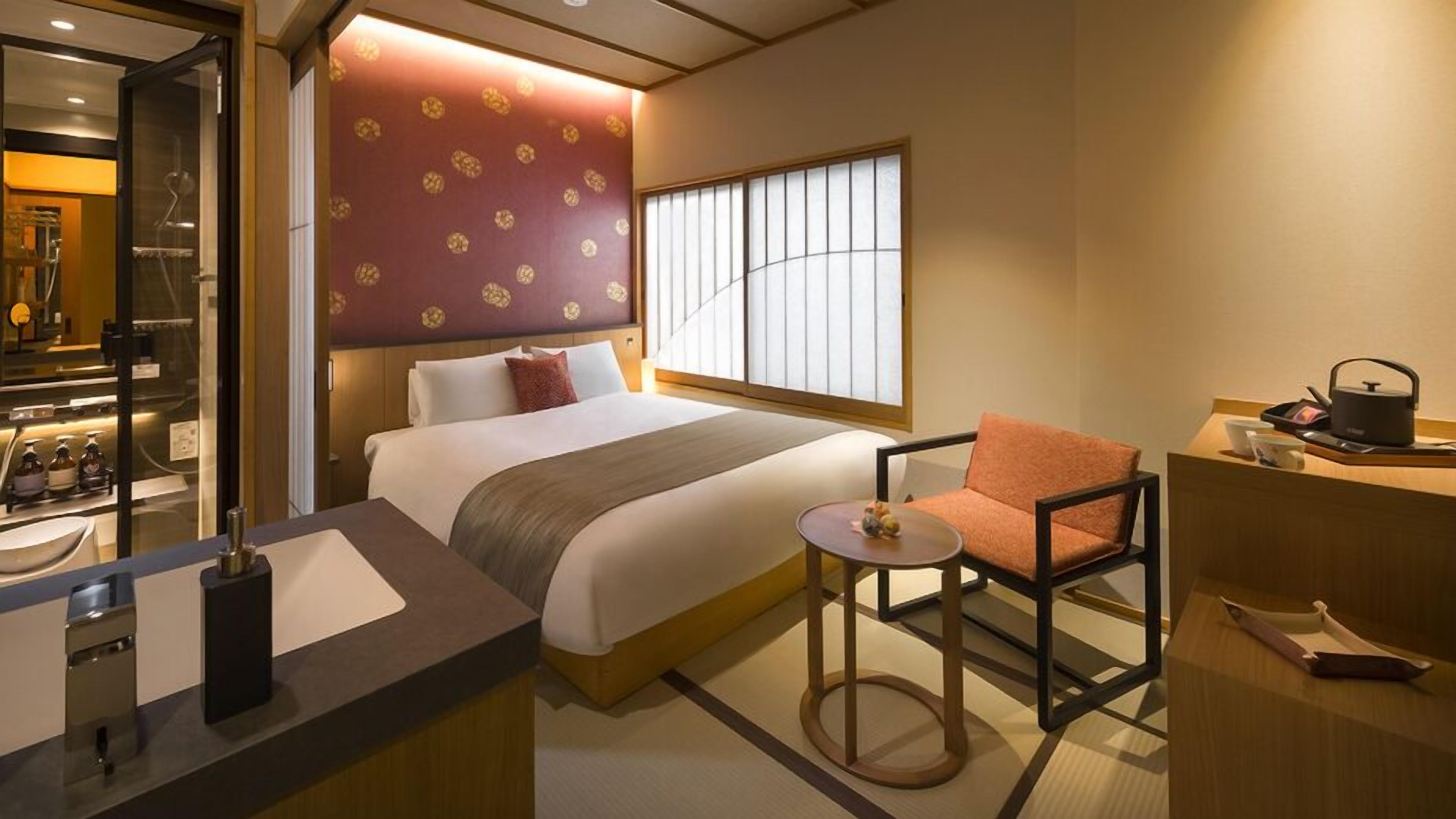 A hotel room with a bed, a chair, a small table, a window, and a kitchenette area.