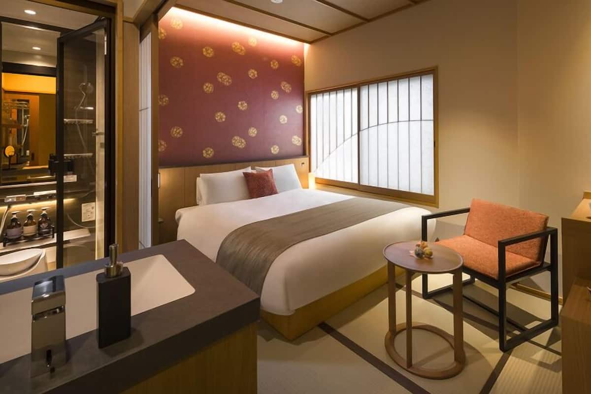 A hotel room with a bed, a chair, a small table, a window, and a kitchenette area.