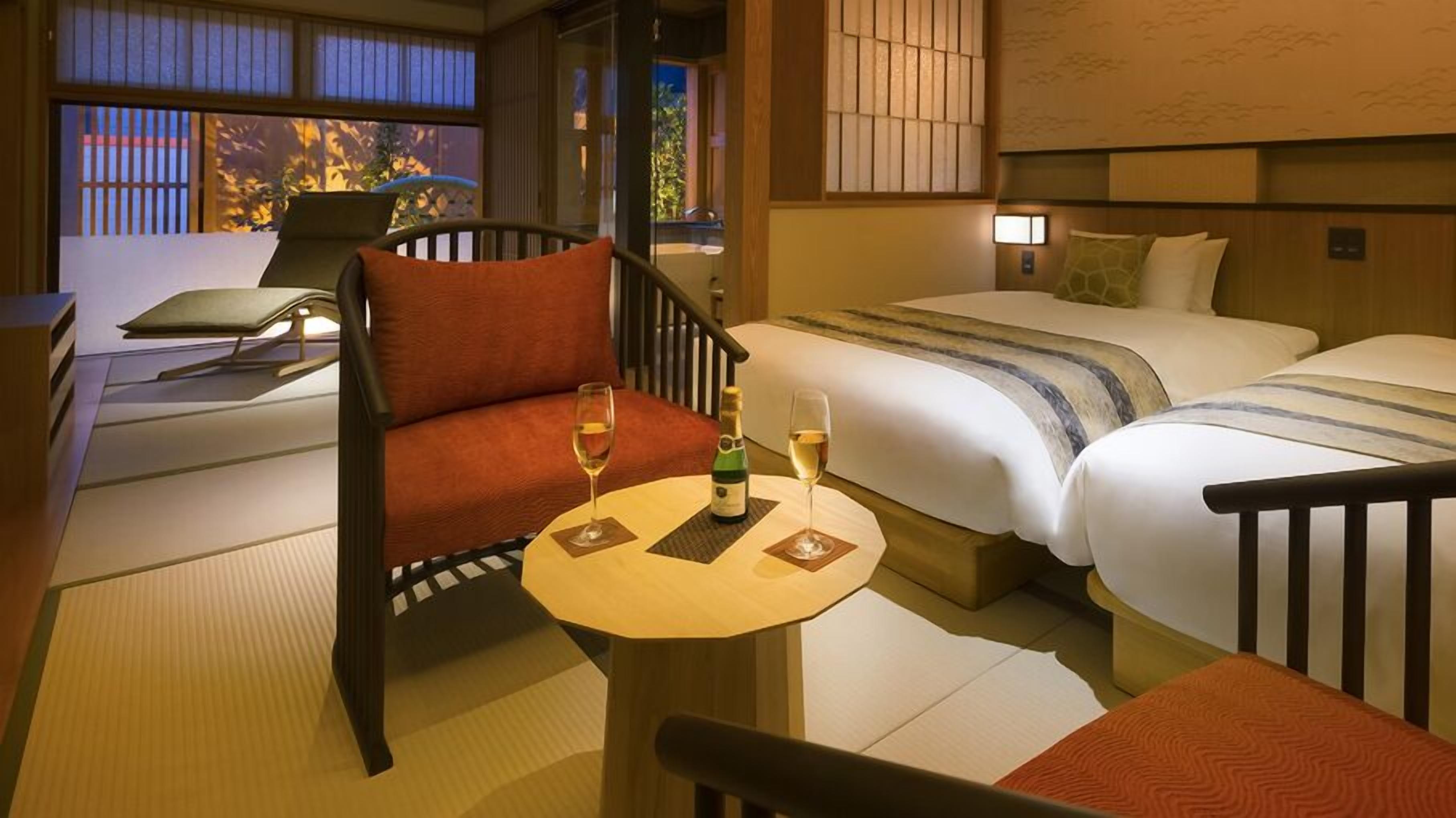 A hotel room with a sofa, a small table with a bottle and glasses, two beds, and a view of the outside.