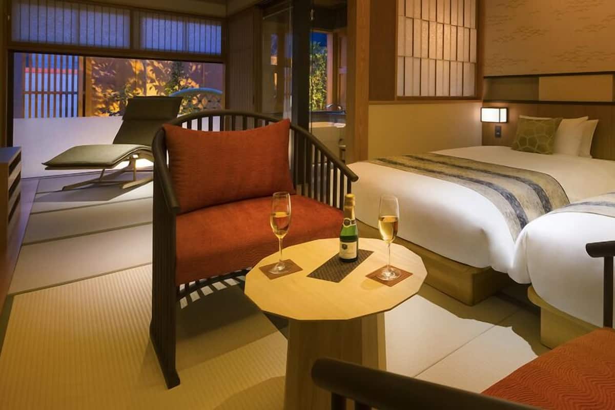 A hotel room with a sofa, a small table with a bottle and glasses, two beds, and a view of the outside.