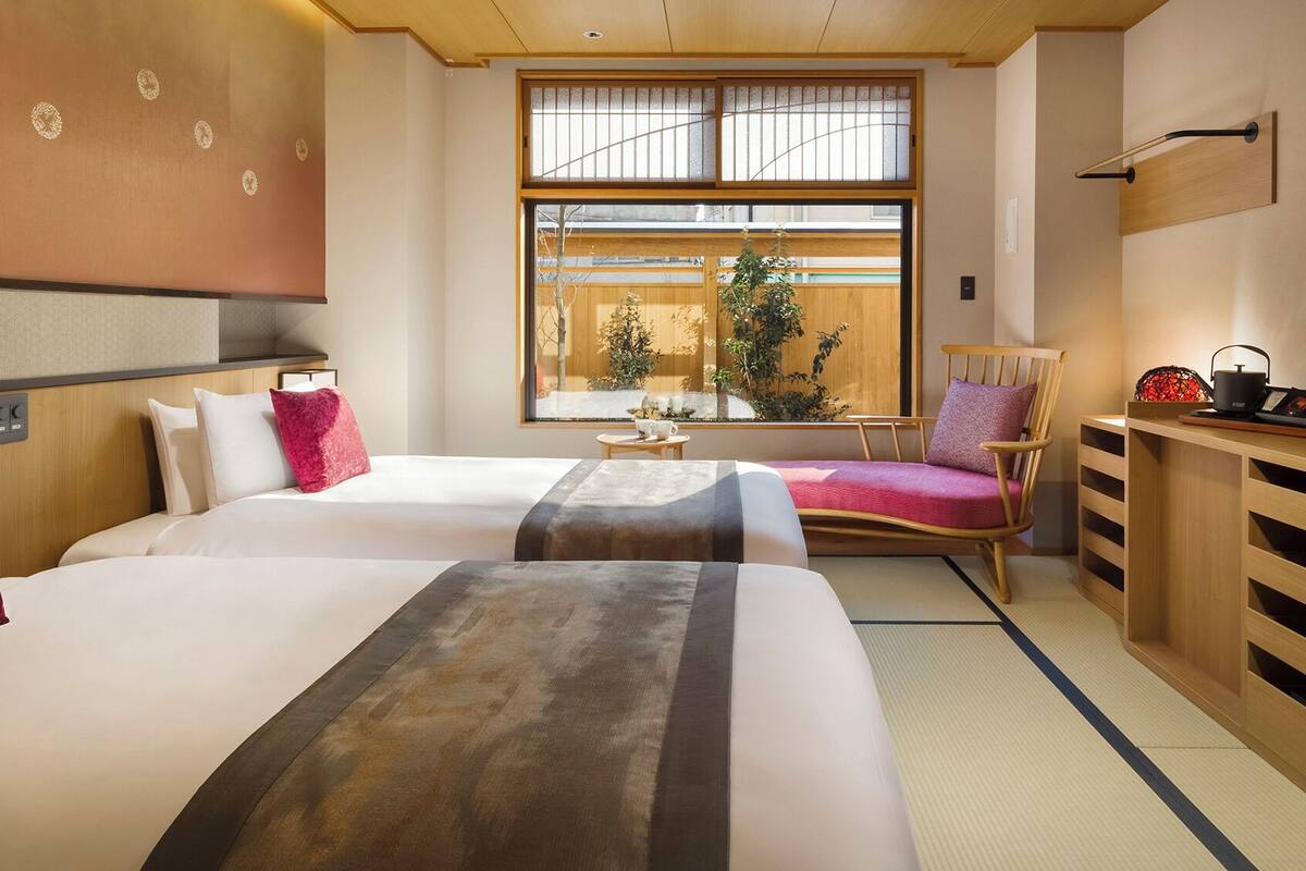 A hotel room with two beds, a TV, a desk with a kettle, and a view of the outside.