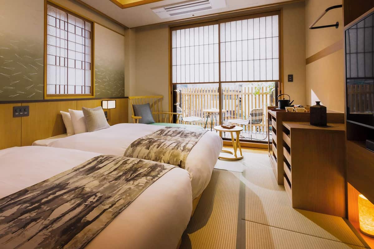 A hotel room with two beds, a desk, a chair, and a view of the outside.